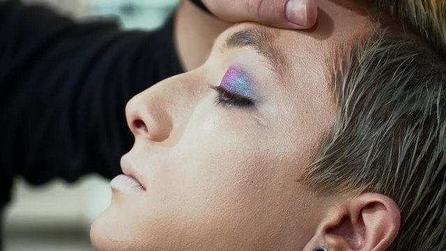 Makeup artist making face art for a young blonde man, painting eyes closeup