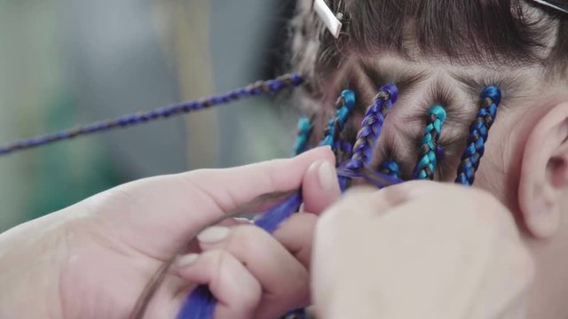 Trend-braids: a modern and topical hairstyle - bright braids. The master plaits African braids with blue artificial strands. Master hairdresser plaits African braids: the process of weaving.