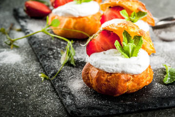 Summer desserts. Homemade baking. Cake profiteroles with whipped cream, fresh strawberries, mint and sprinkling of powdered sugar. On a black stone table. Copy space