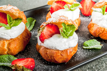 Summer desserts. Homemade baking. Cake profiteroles with whipped cream, fresh strawberries, mint and sprinkling of powdered sugar. On a black stone table. Copy space