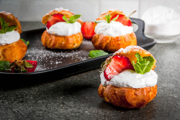 Summer desserts. Homemade baking. Cake profiteroles with whipped cream, fresh strawberries, mint and sprinkling of powdered sugar. On a black stone table. Copy space
