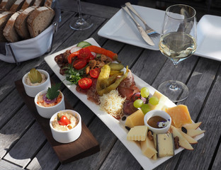 Wooden party table served with glass of white wine and horseradish, various types of cheese, dip, pickles, tomatoes, grape, apple, prosciutto, scratchings and bread.