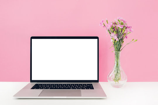 Laptop with white blank screen and flowers in vase on table on pink background. mock up