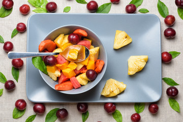 Bowl of fruit salad on rectangular plate. Healthy sliced fresh fruits on a linen grey background. Red berries, green leaves and spoon on table