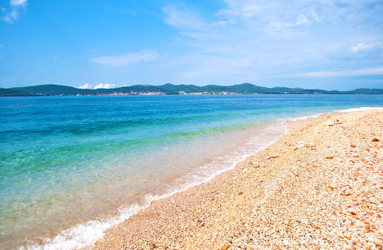 Fototapeta Pebble sandy beach on the background of a hill range with many small houses, a line of buoys and azure transparent blue sea. Blue cloudless sky. Zadar, Croatia