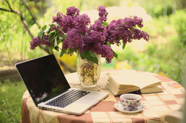 laptop with open book lilac bouquet vase and tea cup on summer garden background 