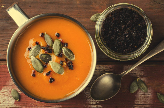 Roasted pumpkin soup in metal mug with seeds and smoked paprika, on rustic wooden background.
