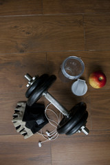 Top view of accessories for fitness in black brown tone. Dumbbells, weight plates, gloves for gym, earphones and bottle for smoothie on wooden background. Concept for sport or workout.