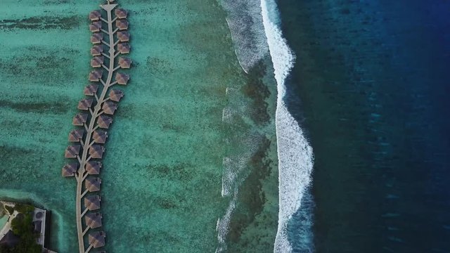 Aerial view of tropical island resort hotel with bungalows and turquoise Indian ocean waves on Maldives, view from above of luxury over water rooms and surf, waves swash