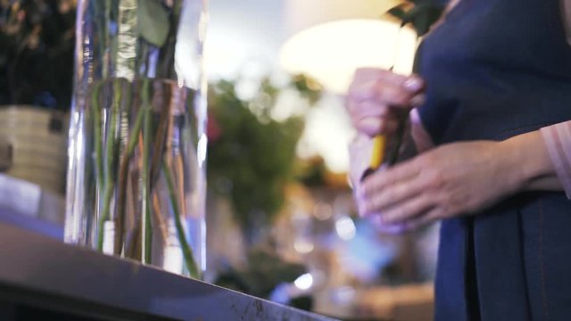 Close up of woman florist cutting rose stems with a knife and putting them inside a glass vase with water. Locked down real time close up shot
