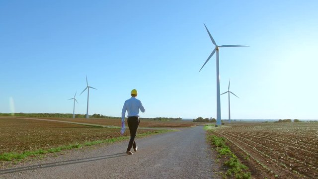 The engineer goes against the background of the windmills, stops and looks at the windmills.