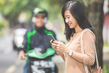 woman standing on sidewalk ordering a commercial motorcycle taxi driver by phone
