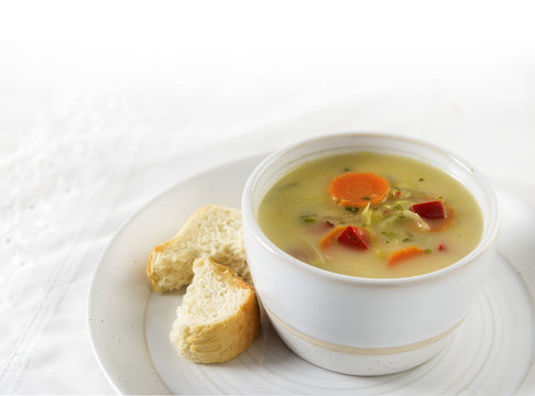 Vegetable cream soup with carrot, red pepper and leek in a bright bowl and bread on a plate, white table cloth, background with copy space fades to white