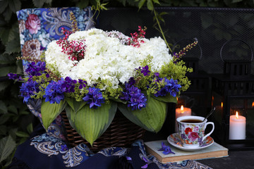 Wicker basket with white hydrangea, blue delphinium, cuff, leaves, hosts in the summer garden. A cup of tea, candles in garden lanterns. Flower composition.