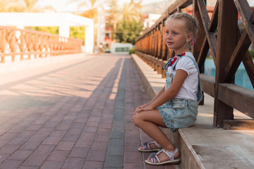 A cute little girl is playfully sitting on a bridge on a summer sunny evening on vacation