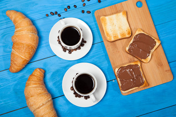 Two cups with coffee, croissant and toast with chocolate on a blue wooden table