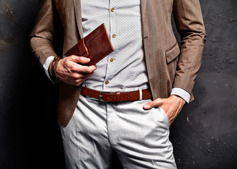 Fashion portrait of young businessman handsome model man dressed in elegant brown suit with accessories on hands posing on gray background in studio. With purse in hands