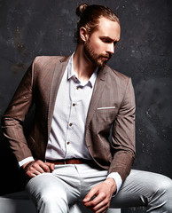 portrait of handsome fashion stylish hipster businessman model dressed in elegant brown suit sitting near dark wall in studio