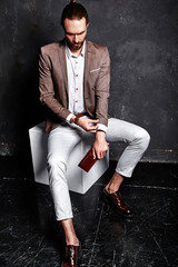 portrait of handsome fashion stylish hipster businessman model dressed in elegant brown suit sitting near dark wall in studio