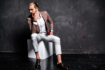 portrait of handsome fashion stylish hipster businessman model dressed in elegant brown suit sitting near dark wall in studio