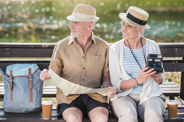 Happy senior couple of travelers sitting together on bench and holding map and instant camera