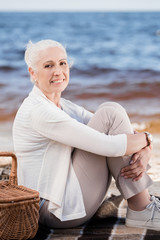 Smiling senior woman sitting on plaid with picnic basket and looking at camera