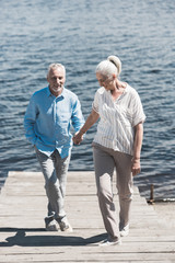 smiling elderly couple holding hands and walking on riverside at sunny day