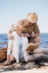 Happy senior man in hat hugging smiling woman sitting on plaid and reading book