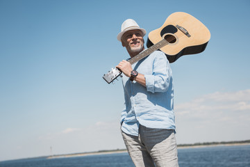 Obraz premium Smiling senior man in hat holding acoustic guitar on shoulder and looking away outdoors