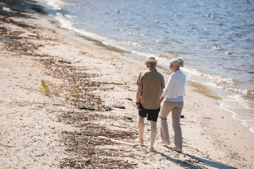 Back view of senior couple holding hands and walking on sand at riverside