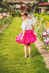 Outdoor portrait of adorable little girl playing in a beautiful flower park on a nice summer day. Fashion kid girl wearing white blouse and bright pink tutu skirt, looking back over shoulder
