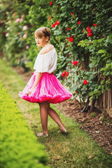 Outdoor portrait of adorable little 9-10 year old little girl wearing white shirt and bright pink tutu skirt, playing in beautiful summer garden