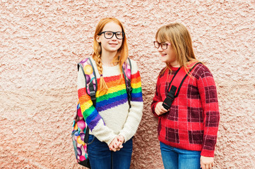 Obraz na płótnie Canvas Group of two adorable kid girls posing outdoors against pink wall, wearing glasses, school backpacks and bright colorful pullovers, back to school concept