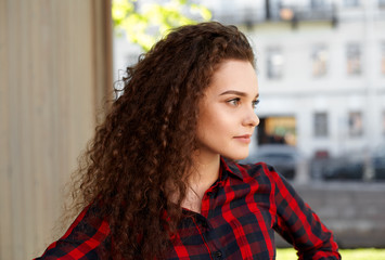 Profile portrait of a beautiful young female with curly hair wearing red plaid shirt with thoughtful and pensive facial expression posing outdoor. Close-up shot of handsome girl on city background