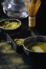 Green tea matcha powder and hot drink in black bowls and wood box standing with glass teapot, bamboo traditional tools spoon, whisk in terracotta tray over dark metal background. Close up