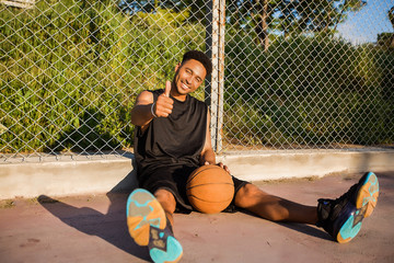 Basketball player is sitting on a basketball court.street ball,sport competitions,afro,outdoor portrait,sport games,handsome black man,pretty,man holding ball,sportsman,black sport shorts,sports shoes