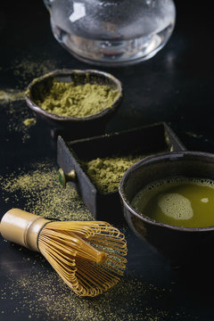 Green tea matcha powder and hot drink in black bowls and wood box standing with glass teapot, bamboo traditional tools spoon, whisk in terracotta tray over dark metal background. Close up