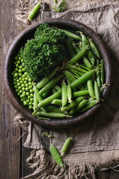 Young organic green pea pods and peas in terracotta tray and bundle of parsley over old dark wooden planks with sackcloth textile background. Top view with space. Harvest, healthy eating.
