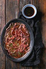 Asian style Beef carpaccio with soy sauce, chive onion, black sesame, served on vintage metal tray with textile, bowl of sauce over old wooden plank background. Top view with space