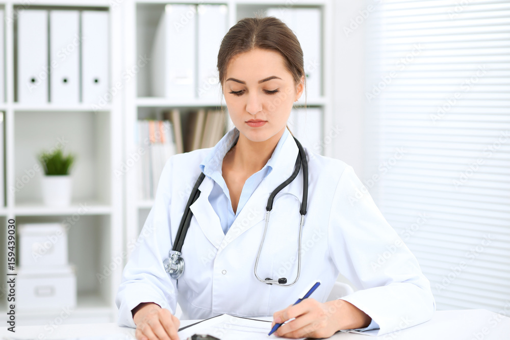 Poster young brunette female doctor sitting at the table and working at hospital office.