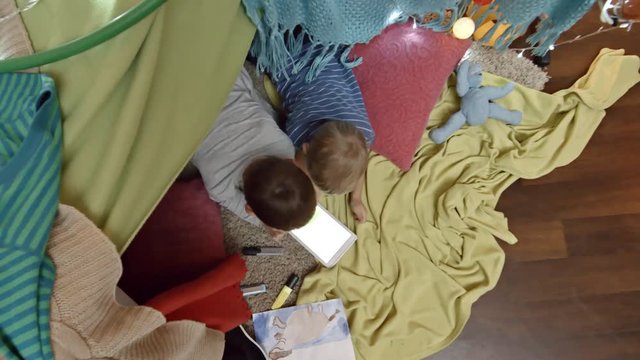 Zoom in shot of two little brothers lying on floor under play tent and using digital tablet. View from above of drawing, felt tip pens and toys surrounding boys