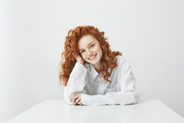 Beautiful ginger girl with curly hair smiling looking at camera sitting at table over white background.