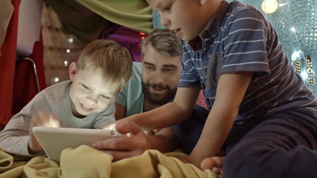 Tracking shot of father and two little sons lying on carpet under play tent decorated with lights and playing game on digital tablet
