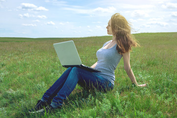 Woman sitting on a green meadow on the background of sky with clouds and working or studying with laptop wireless