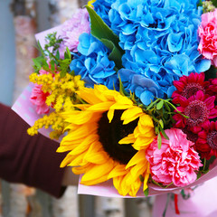 The florist holds in hands a beautiful bright summer bouquet of flowers on the background of wooden trunks. Blue hydrangea, sunflower, pink carnation, solidago
