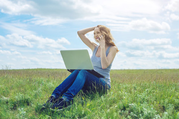 Naklejka na ściany i meble Woman sitting on a green meadow on the background of sky with clouds and working or studying with laptop wireless