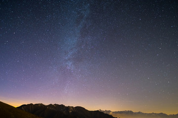 Fototapeta na wymiar The wonderful starry sky on Christmas time and the majestic high mountain range of the Italian French Alps, with glowing villages below and moonlight.