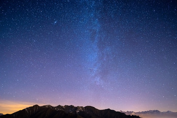 Fototapeta premium The wonderful starry sky on Christmas time and the majestic high mountain range of the Italian French Alps, with glowing villages below and moonlight.