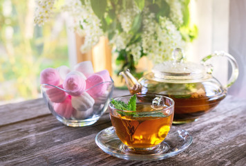 Mint tea in glass Cup on the background of a vase with marshmallows and a transparent glass teapot in a Sunny summer morning.