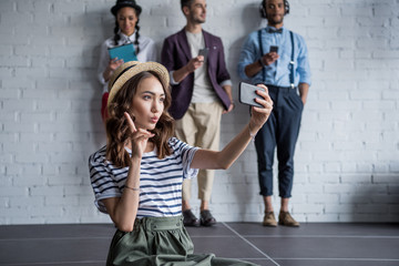 young stylish woman taking selfie on smartphone while her friends using digital devices